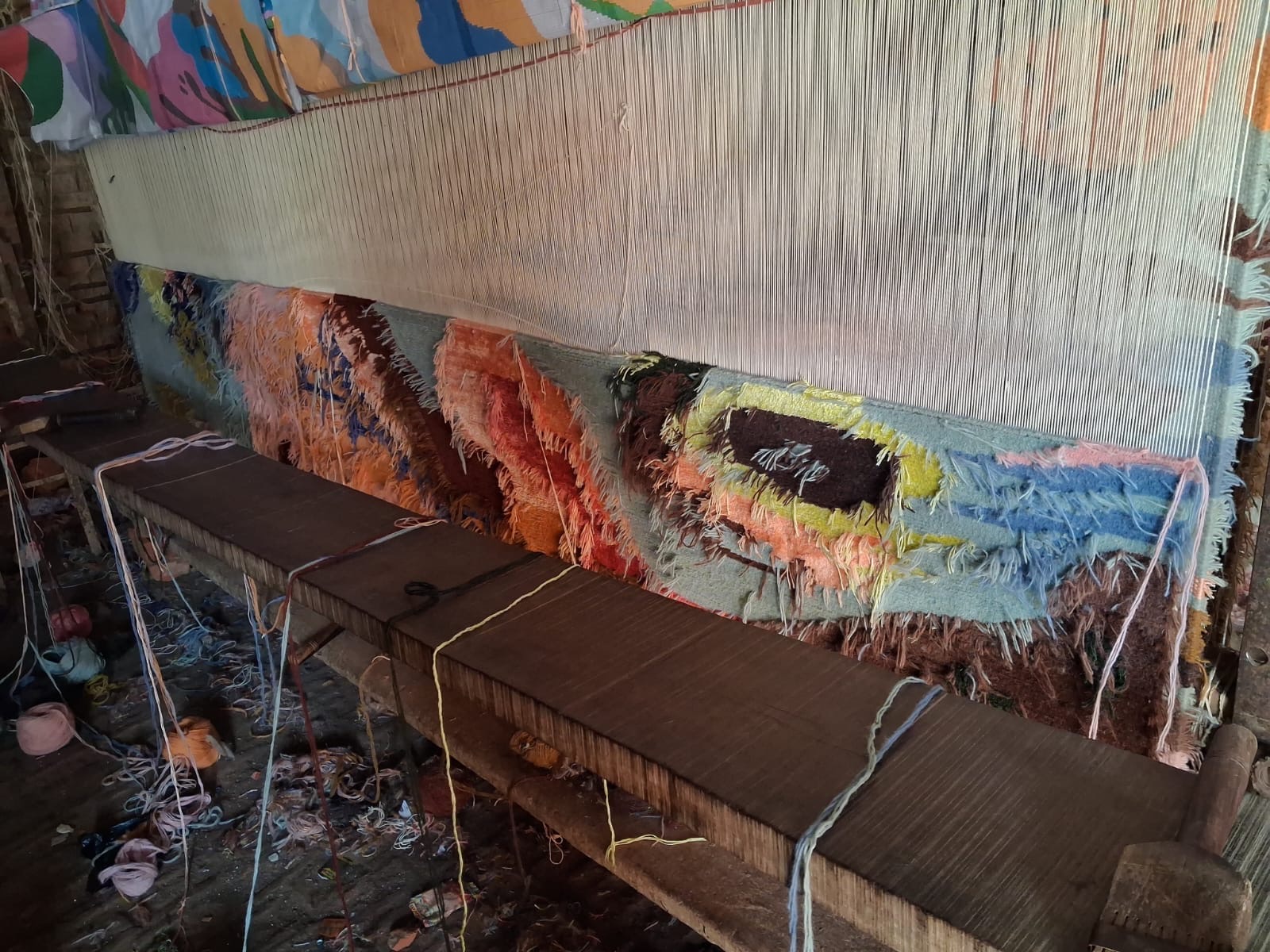 The carpet on the loom during weaving, with the painted colour plan hanging above